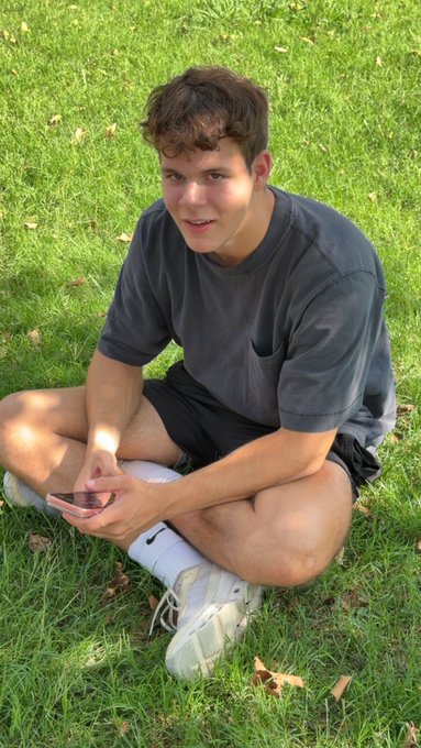 A young man with curly brown hair sits cross-legged on green grass, wearing a dark gray t-shirt, black shorts, and white sneakers while holding a phone. The casual outdoor setting matches the laid-back tone of the post about creating "$ANSEM content with mega neck @ElectroExtant," suggesting a relaxed, behind-the-scenes moment.