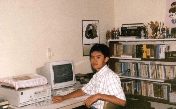 A young man sits at a computer in a room filled with bookshelves and a vintage boombox, with a framed poster on the wall behind him.