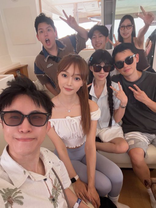 A group selfie of seven people posing playfully on a boat, with several making peace signs and goofy faces, wearing casual summer outfits and sunglasses.