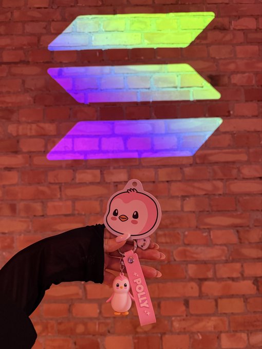 A hand holds a pink "POLLY" keychain featuring a cartoon penguin and owl against a brick wall illuminated by colorful neon arrow lights, tying into the "Pengu Solana morning & gm" post with its cute crypto-themed mascot charm.