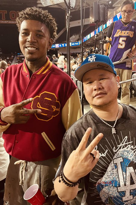Two men pose at what appears to be an LA Lakers game, with one wearing a red and yellow "SC" varsity jacket and the other in an LA Dodgers cap and Air Jordan shirt, while a Lakers fan (#5 jersey) stands in the background holding a sign. The image supports the post's caption of a casual networking encounter at a Lakers game, where the man in the SC jacket is being pitched on the $APE token.