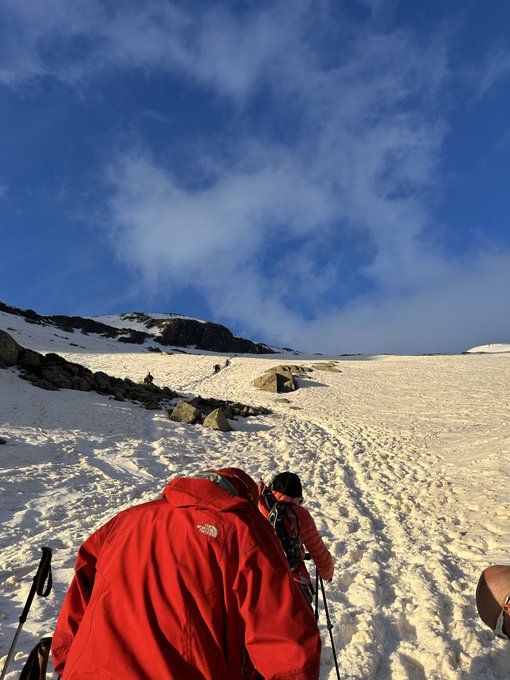 Two hikers in winter gear, one wearing a red North Face jacket, ascending a snow-covered mountain trail under a bright blue sky with wispy clouds.