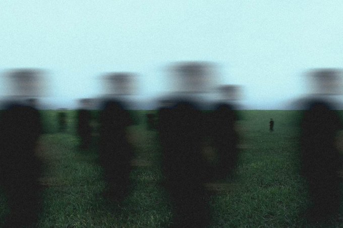 A blurry, low-resolution image showing dark silhouetted figures standing in a grassy field beneath a hazy, overcast sky.