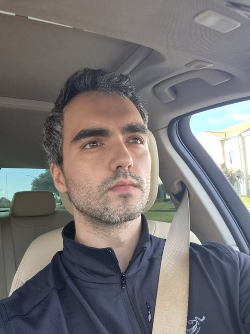 A man with dark hair and a short beard sits in the driver's seat of a car, wearing a dark zip-up jacket and secured with a seatbelt, gazing thoughtfully out the window as he travels—capturing the focused, on-the-move mindset of heading to support the next big project on @MetaDAOProject.