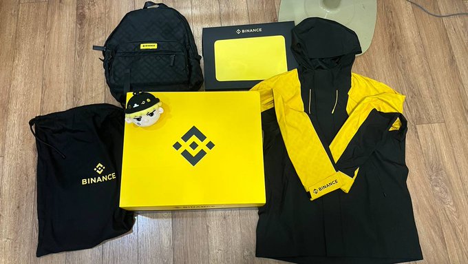 A flat lay of Binance-branded merchandise on a wooden floor, including a yellow and black jacket, backpack, drawstring bags, and a yellow box featuring the Binance logo.