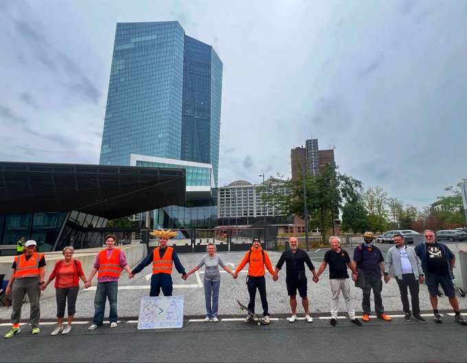 A group of people including activists in orange vests hold hands forming a human chain in protest on a city street with modern skyscrapers in the background.