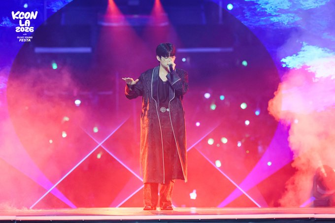 A performer in a long coat sings on stage amid vibrant pink and blue lighting at KCON LA 2026, with "KCON LA 2026 OLIVE YOUNG FESTA" branding visible in the top-left corner, capturing a moment from the M COUNTDOWN stage at the Crypto.com Arena event.
