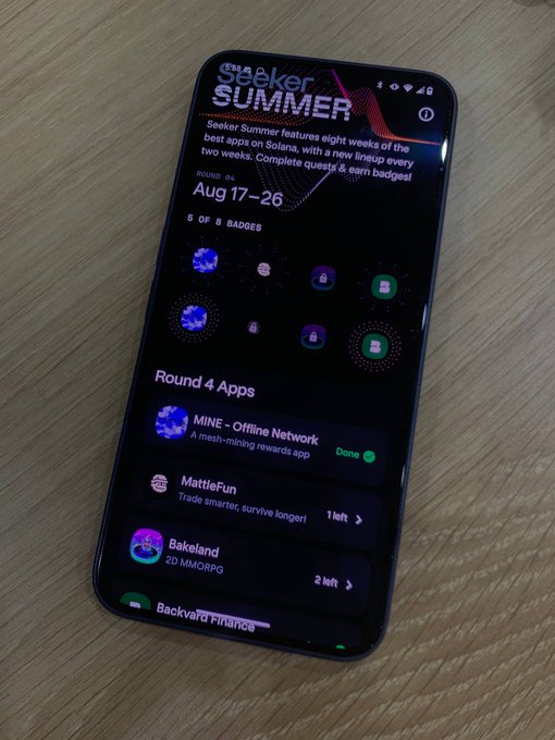 A smartphone resting on a wooden surface displays a "Seeker Summer" event page (Round 04, Aug 17-26) with a list of apps to complete quests for, including "MINE - Offline Network" (marked Done), "MattleFun," and "Bakeland."