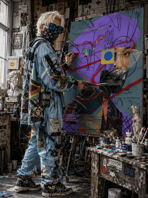 An artist in blue overalls covered in stickers and graffiti stands in a chaotic studio, painting an abstract mixed-media portrait featuring a gas-masked figure with colorful shapes and text including 'WAGMI' and 'R3DRDR'.