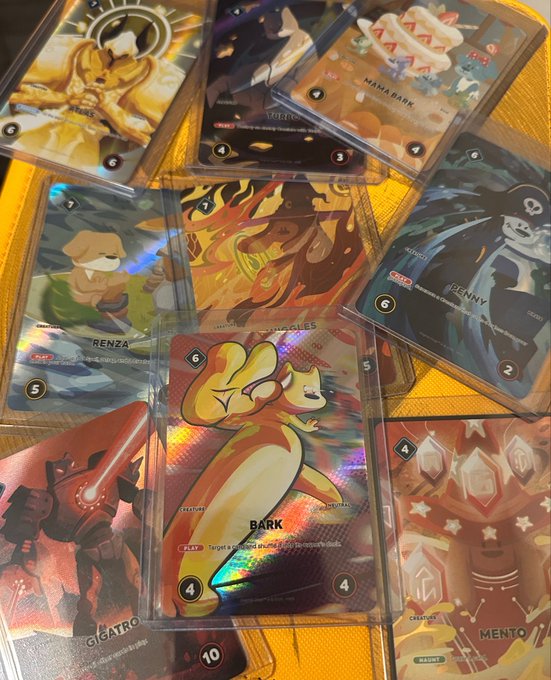A collection of trading card game cards featuring characters like BARK, RENZA, MANA BARK, GIGATRON, and MENTO spread on a wooden surface, with BARK's holographic card prominently displayed showing stats of 4/4 and 'Target is removed after the duel.'