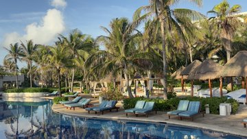 A tropical resort scene featuring palm trees, thatched-roof structures, a swimming pool, and lounge chairs under a sunny sky.
