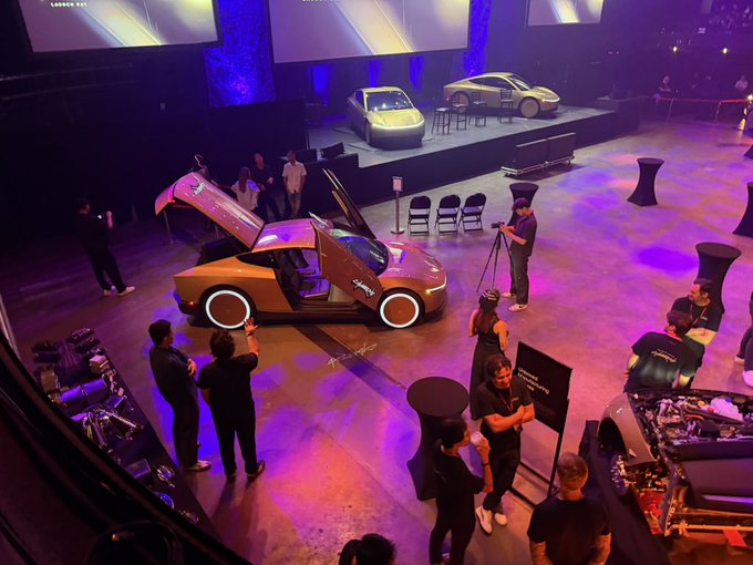 An elevated view of an indoor car exhibition featuring futuristic vehicles on display platforms, with attendees and photographers gathered around a gold-colored car with illuminated wheel rims.