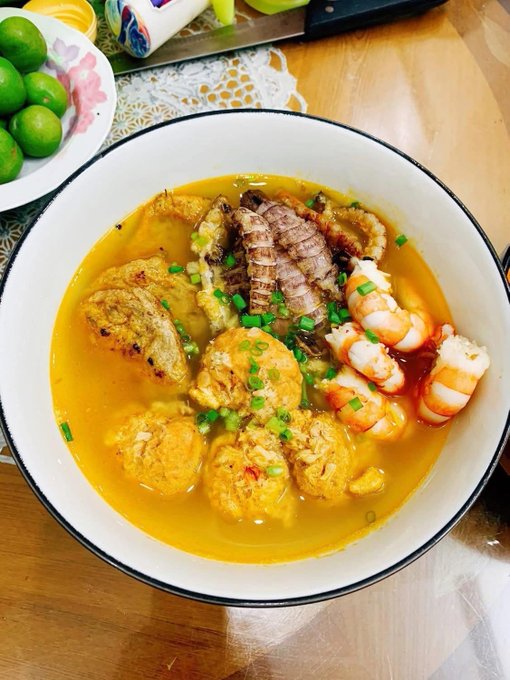 A bowl of seafood soup contains shrimp, lobster tail, and fried items in a rich yellow broth, garnished with green onions and served on a wooden table with limes nearby.
