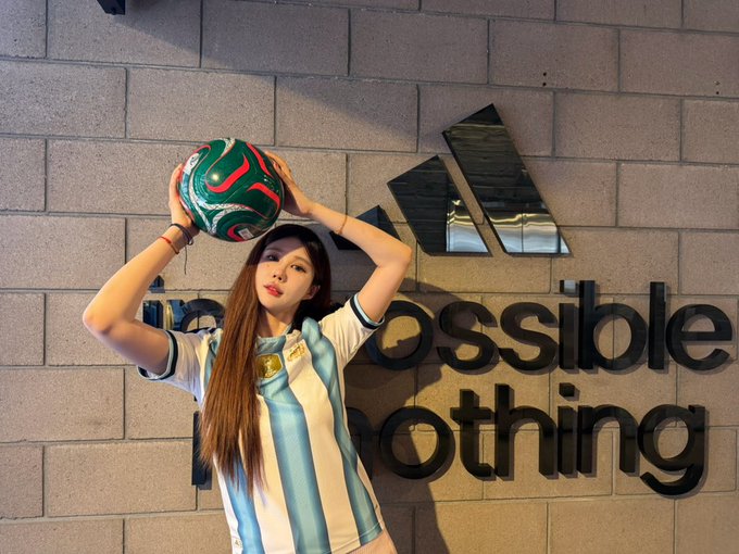 A woman wearing an Argentina national football team jersey holding a soccer ball above her head while posing in front of an Adidas 'Impossible is Nothing' branded wall.