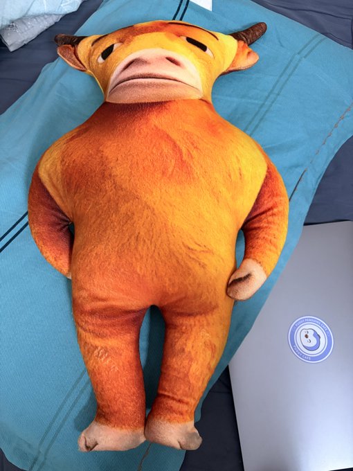 An orange plush cow with horns and a pink snout lies on a blue cushion next to a laptop, matching the post's humorous description of an ugly stuffed cow sent for crypto "luck," with the "888" number visible in the caption referencing the lucky number superstition.