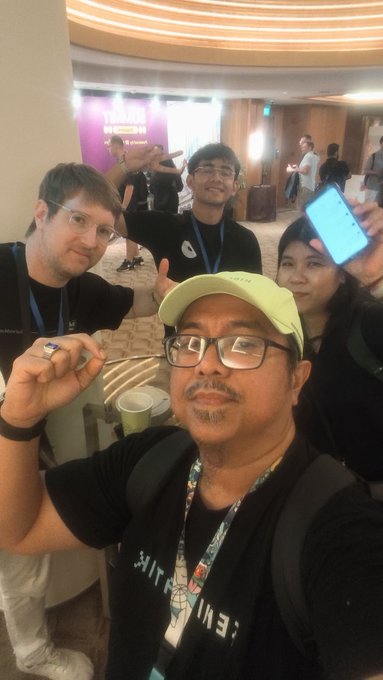 A group of people pose for a selfie at what appears to be a conference, with one man in the foreground making a hand gesture and wearing a lanyard.