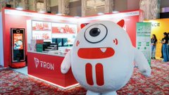 A small white monster mascot with one eye and fangs posing in front of a 'TRON' branded booth at what appears to be a convention or event.
