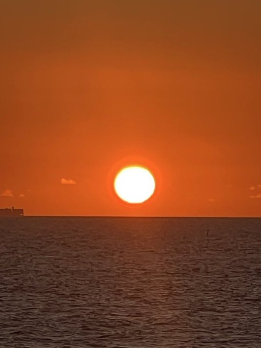 A bright white sun setting over a calm ocean, casting an orange glow across the sky and water.