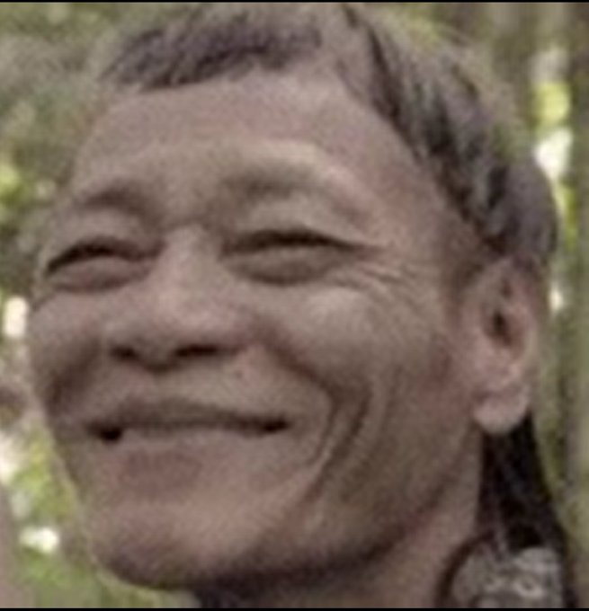 A close-up of a person with short dark hair smiling broadly outdoors, with green foliage visible in the blurred background.