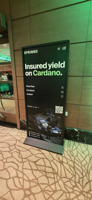 A digital advertising display reading 'PRIME | α: Insured yield on Cardano. Fixed Rate, Compliant, Audited' with a QR code, displayed on a patterned carpet.