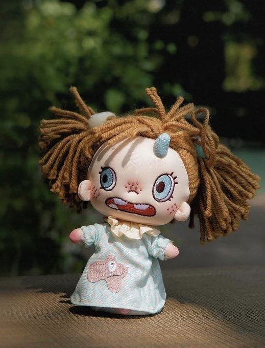 A handmade cloth doll with yarn hair, large embroidered eyes, and a stitched mouth wearing a light blue dress with a pink animal appliqué.
