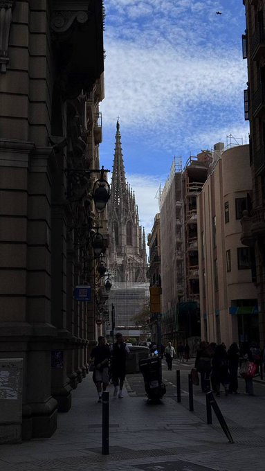 A narrow Barcelona street frames a striking view of the spires of the Sagrada Família rising against a partly cloudy sky, capturing the architectural marvel that defines the city. The scene reflects the post's highlight of experiencing Barcelona's iconic sights during a spring visit to Spain before heading to ETHCC.
