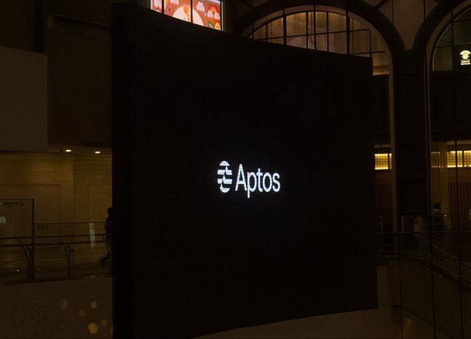 An illuminated 'Aptos' logo is displayed on a dark panel in a building interior at night, with arched windows visible in the background.