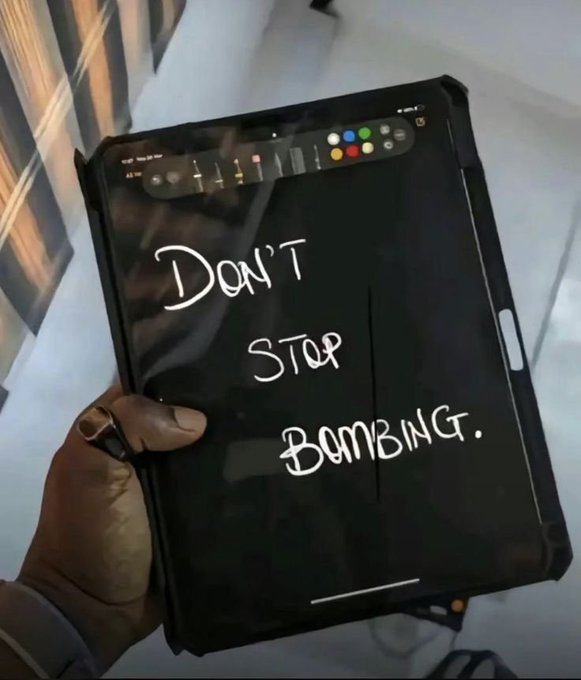 A person holding a tablet with a black screen displaying the handwritten message 'DON'T STOP BOMBING.' in white text.
