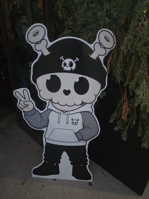 A cardboard cutout of the cartoon character Kuromi from Sanrio stands outdoors, flashing a peace sign against a backdrop of greenery and pavement, visually representing the staking of $ETH with the Quirky NFT project.