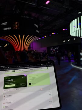 A laptop on a table shows a webpage with $156.86, while a colorful event with people and a large illuminated installation can be seen in the background.