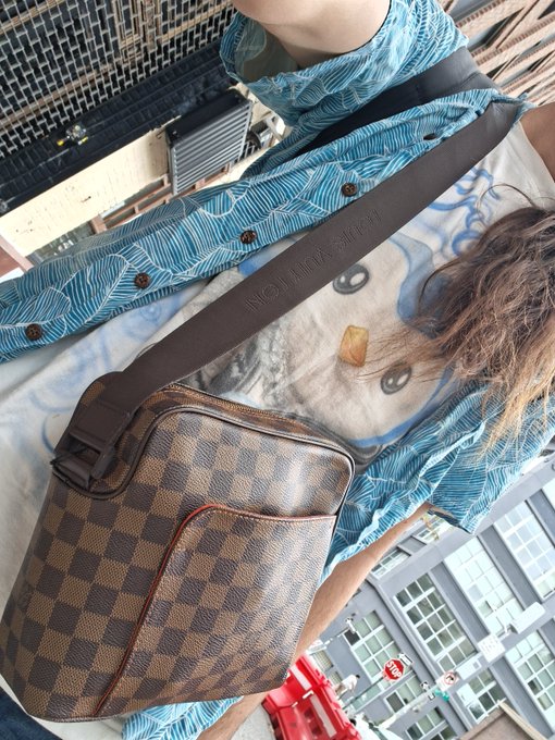 A person wearing a blue patterned shirt and a white graphic t-shirt carries a brown Louis Vuitton Damier Ebene messenger bag across their body in an urban setting.