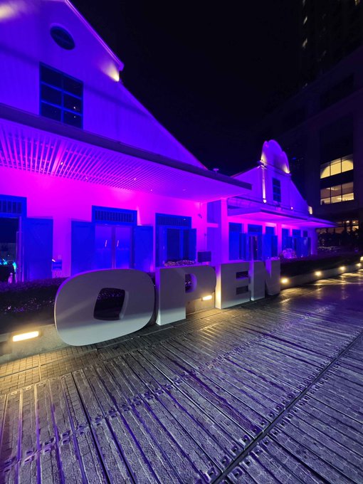 A building exterior is illuminated in purple and pink light at night, with large white 'OPEN' letters sitting on a wooden deck.