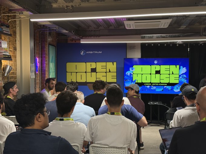 A photograph of an 'Arbitrum Open House' event in London, United Kingdom, showing an audience seated facing two large screens displaying the event's logo.