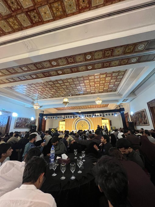 A large indoor gathering of attendees seated at round tables in an ornate hall with decorative ceiling patterns, facing a stage with a large screen and golden logo during what appears to be a conference or seminar.