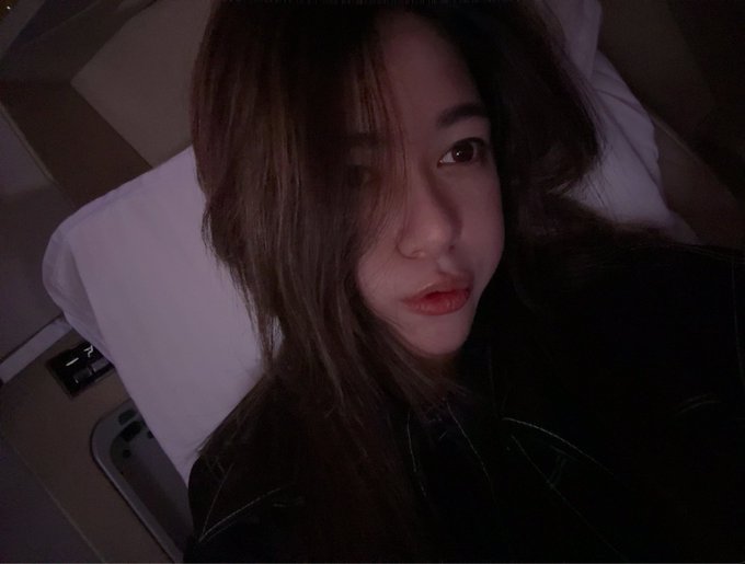 A selfie of a young woman with long brown hair in a dimly lit setting, possibly on a train or bus.