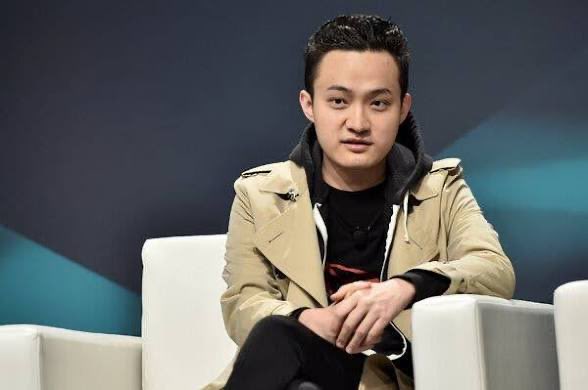 A young man with dark hair wearing a beige trench coat over a dark shirt sits in a white armchair, appearing to be on a panel or interview with a dark background.