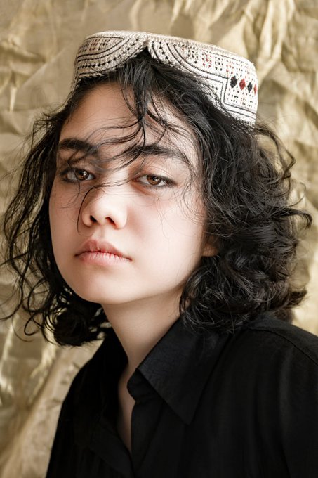 A portrait of a person with dark curly hair wearing a traditional patterned cap and a black collared shirt, posed against a textured gold backdrop.