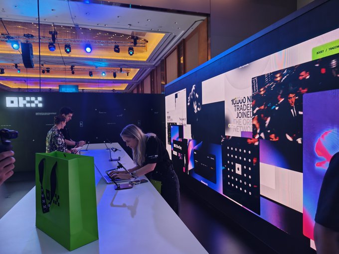 A trade show booth with an 'OKX' logo where staff assist attendees at a counter next to large display screens.