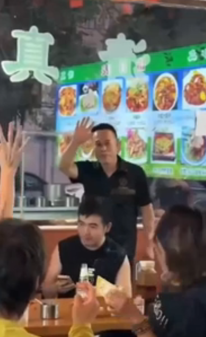 A restaurant scene with a man in a black shirt waving at patrons, with Chinese menu boards visible in the background showing various dishes.