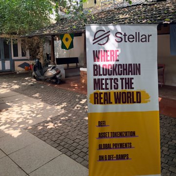 A yellow and white Stellar banner reading 'WHERE BLOCKCHAIN MEETS THE REAL WORLD' with features listed including DeFi, asset tokenization, global payments, and on/off-ramps, displayed outdoors near a building.