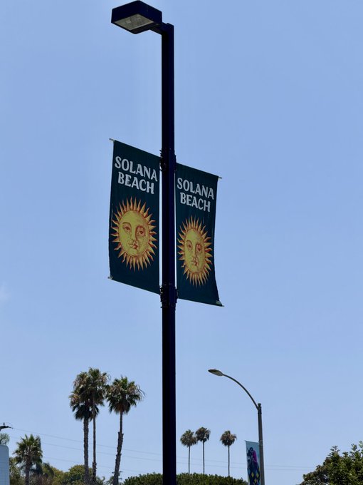 A street lamp displays two 'Solana Beach' banners featuring a sun face logo against a clear blue sky, with palm trees visible below.