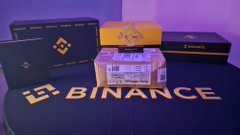 A Binance promotional display features branded merchandise including boxes, a notebook, and a candle arranged around the Binance logo.