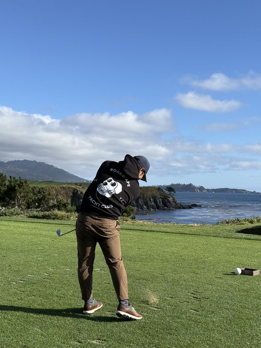 A person in a black hoodie and cap swinging a golf club on a coastal course with ocean and mountains visible in the background.