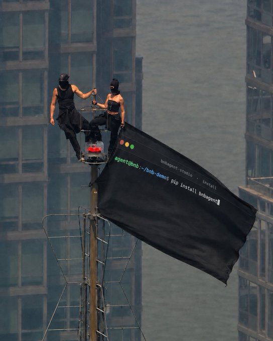 Two masked performers on top of a structure holding a large black banner styled like a terminal window with command-line text promoting "bnbagent-studio" via pip install.
