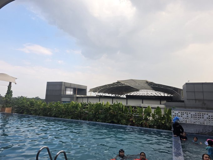 A rooftop infinity pool view overlooking a stadium-like building, with several people in the water including children.