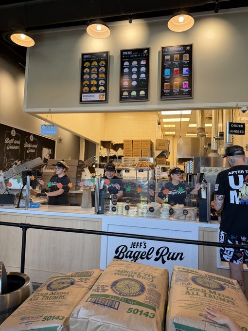 Staff at "Jeff's Bagel Run" prepare orders behind the counter, with digital menu boards displaying bagels, spreads, and featured drinks overhead, while large flour sacks labeled "Gold Medal All Trumps" (50143) are stacked in the foreground. The bustling bagel shop scene fits the caption's casual Sunday morning vibe of grabbing coffee and bagels, contrasting with the celebratory tone of a profitable $SUI trade.