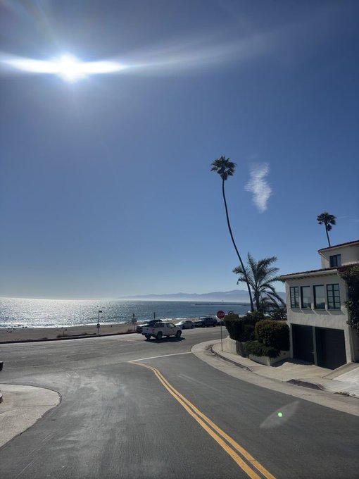 A sunny coastal road curves toward the ocean, with palm trees, parked cars, and a Mediterranean-style house on the right under a bright sun.