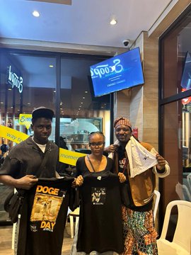 iced coffee shop with a large window displaying "Good morning" and "DOGE DAY" signs, accompanied by two individuals holding black t-shirts.