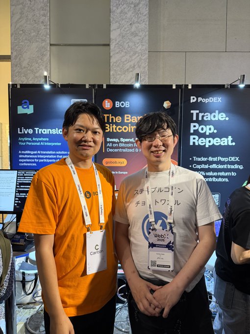 Two men smiling and posing together at a Web3 conference, standing in front of booths with signage for BOB ('The Bank of Bitcoin', gobob.xyz), PopDEX ('Trade. Pop. Repeat.'), and a 'Live Translation' service.