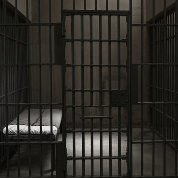 A dark, foreboding image of an empty prison cell with iron bars, a simple cot, and a toilet, symbolizing the potential lengthy incarceration faced by the Brooklyn man sentenced to up to 12 years for stealing $16 million from Coinbase users through a phishing scam.